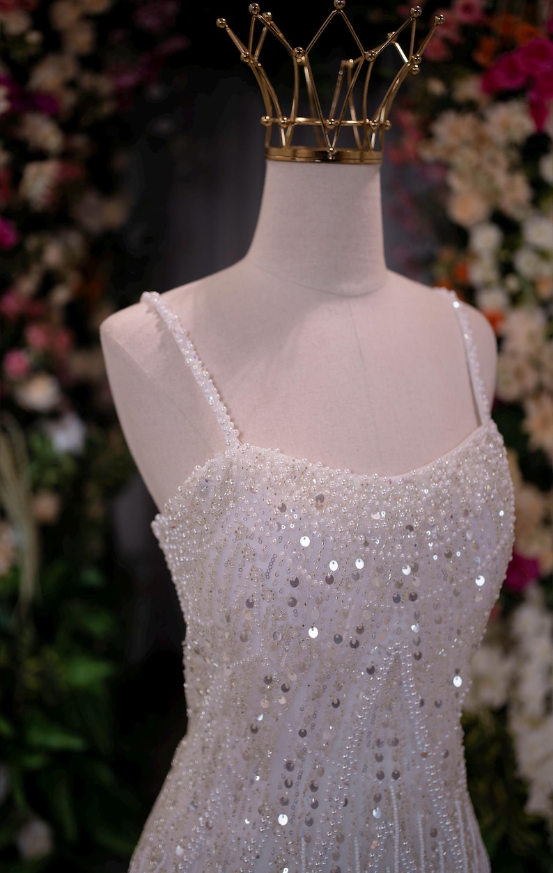 May include: A white, beaded wedding dress with a sweetheart neckline and spaghetti straps. The dress is adorned with sequins and small beads. A gold crown sits atop a mannequin head, suggesting a bridal theme.