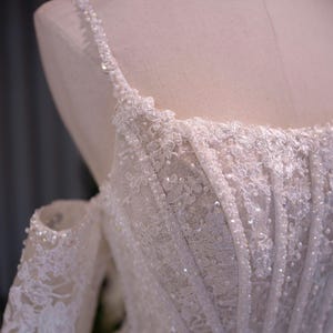 K&ouml;nnte beinhalten: Nahaufnahme eines wei&szlig;en Brautkleid-Oberteils auf einer Schaufenpuppe. Das Kleid weist aufwendige Spitzendetails, glitzernde Pailletten und zarte, mit Perlen besetzte Tr&auml;ger auf. Das schulterfreie Design und die detaillierten Blumenmuster schaffen eine elegante und romantische &Auml;sthetik.