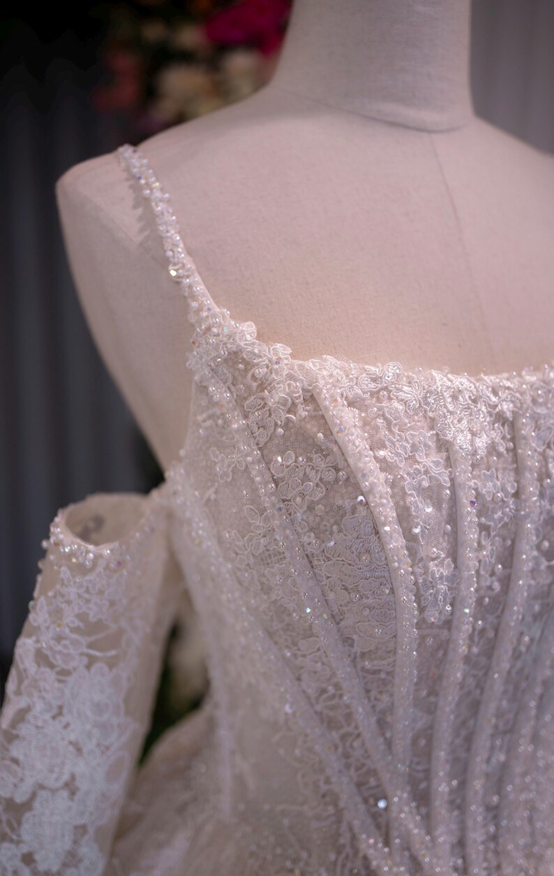 K&ouml;nnte beinhalten: Nahaufnahme eines wei&szlig;en Brautkleid-Oberteils auf einer Schaufenpuppe. Das Kleid weist aufwendige Spitzendetails, glitzernde Pailletten und zarte, mit Perlen besetzte Tr&auml;ger auf. Das schulterfreie Design und die detaillierten Blumenmuster schaffen eine elegante und romantische &Auml;sthetik.