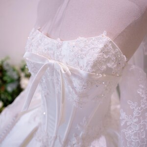 May include: A white strapless gown featuring a corset-style bodice with vertical boning, intricate floral lace embroidery, and scattered iridescent sequins. A thin satin ribbon bow is tied at the center chest, and sheer lace sleeves extend outward.
