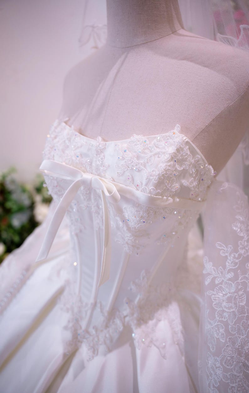 May include: A white strapless gown featuring a corset-style bodice with vertical boning, intricate floral lace embroidery, and scattered iridescent sequins. A thin satin ribbon bow is tied at the center chest, and sheer lace sleeves extend outward.