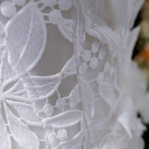May include: Close-up of white lace fabric with floral and leaf patterns. The delicate lace features intricate embroidery and a soft, textured appearance. The design includes small, beaded accents and layered fabric details.