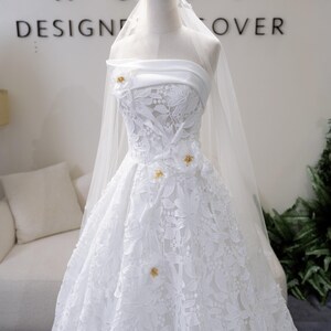 May include: A white bridal gown with intricate floral lace and a flowing skirt, displayed on a mannequin. The dress features a sheer veil and small gold embellishments. The word "ROMY" is visible on the wall behind the dress.