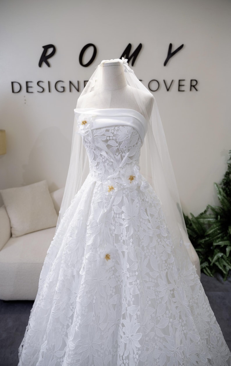 May include: A white bridal gown with intricate floral lace and a flowing skirt, displayed on a mannequin. The dress features a sheer veil and small gold embellishments. The word "ROMY" is visible on the wall behind the dress.