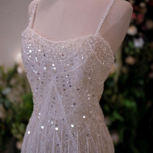May include: A white, beaded formal gown with spaghetti straps. The bodice is heavily embellished with sequins and beads, and the skirt features vertical beaded fringe. The dress is displayed on a mannequin.