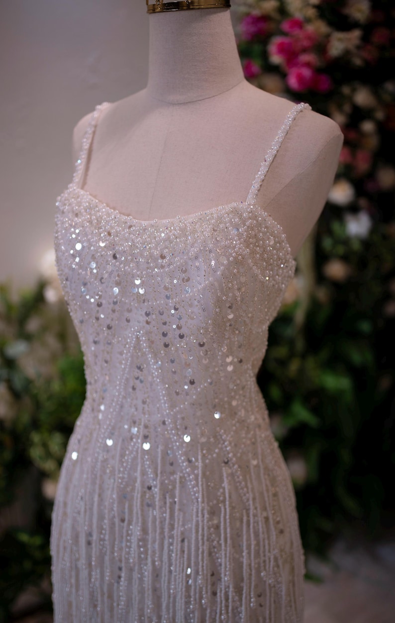 May include: A white, beaded formal gown with spaghetti straps. The bodice is heavily embellished with sequins and beads, and the skirt features vertical beaded fringe. The dress is displayed on a mannequin.