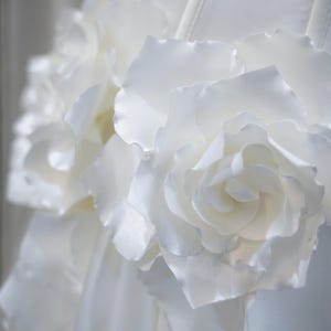 May include: A white garment featuring large, layered fabric rose embellishments. The flowers are constructed from smooth, matte white material with delicate, curved petals that create a dimensional, textured surface across the front of the piece.
