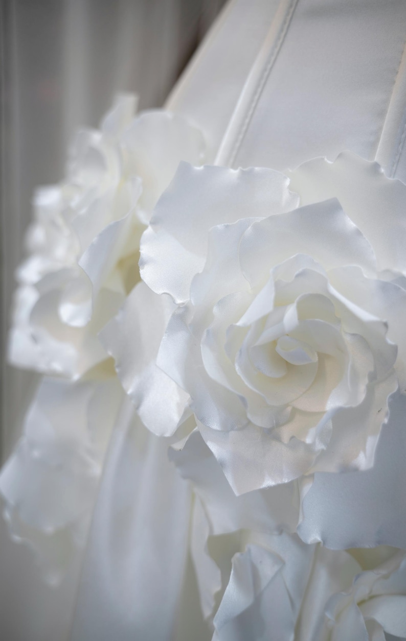 May include: A white garment featuring large, layered fabric rose embellishments. The flowers are constructed from smooth, matte white material with delicate, curved petals that create a dimensional, textured surface across the front of the piece.