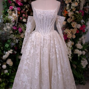 K&ouml;nnte beinhalten: Wei&szlig;es Hochzeitskleid mit eng anliegendem Oberteil und weitem Rock, ausgestellt auf einer Schaufensterpuppe. Das Kleid hat aufwendige Spitzendetails und schulterfreie &Auml;rmel. Auf dem Kopf der Schaufensterpuppe sitzt eine goldene Krone. Der Hintergrund ist ein Blumenarrangement.