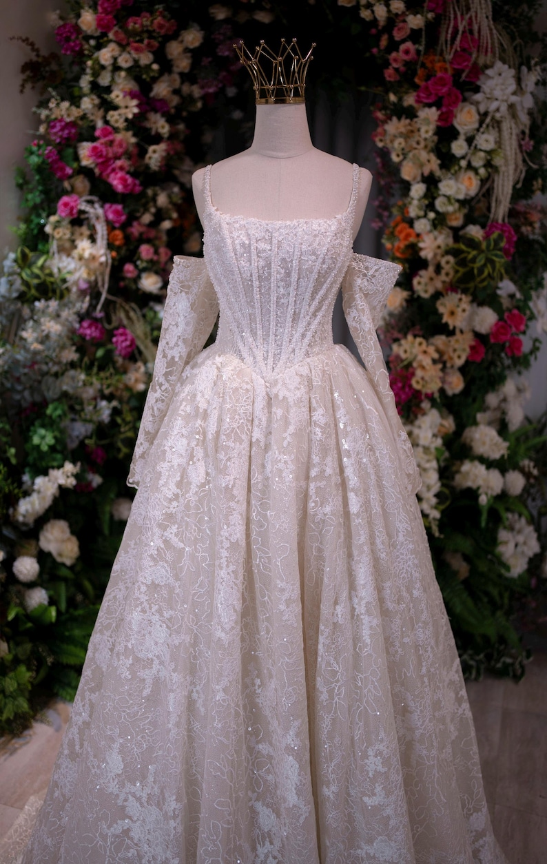 K&ouml;nnte beinhalten: Wei&szlig;es Hochzeitskleid mit eng anliegendem Oberteil und weitem Rock, ausgestellt auf einer Schaufensterpuppe. Das Kleid hat aufwendige Spitzendetails und schulterfreie &Auml;rmel. Auf dem Kopf der Schaufensterpuppe sitzt eine goldene Krone. Der Hintergrund ist ein Blumenarrangement.