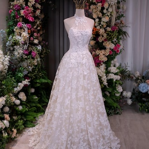 May include: A floor-length white gown featuring a high halter neckline with intricate lace floral appliqu&eacute;s over a sheer bodice. The full skirt is layered with white lace embroidery over a neutral-toned base, creating a structured silhouette.