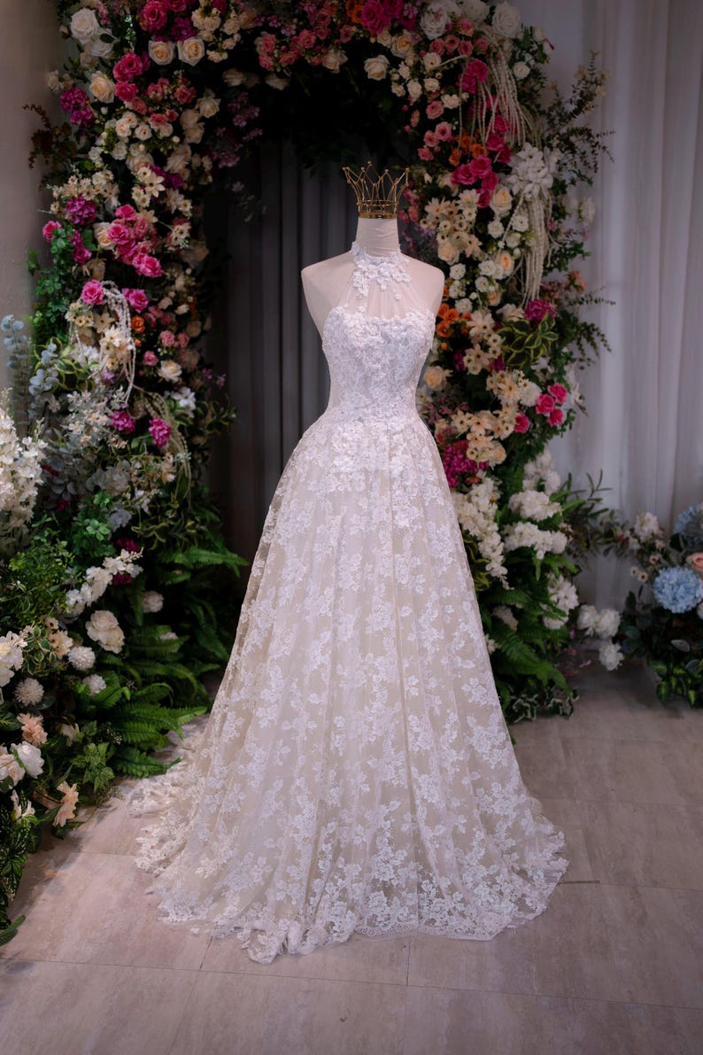 May include: A floor-length white gown featuring a high halter neckline with intricate lace floral appliqu&eacute;s over a sheer bodice. The full skirt is layered with white lace embroidery over a neutral-toned base, creating a structured silhouette.