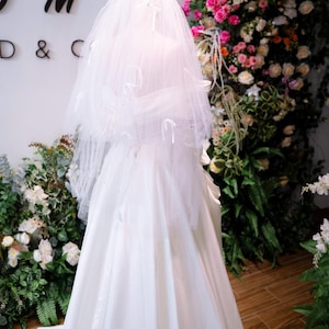 May include: A white wedding dress and veil on a mannequin. The veil is adorned with subtle decorations. The dress has a flowing skirt. The background includes floral arrangements with pink and white flowers, and green foliage.