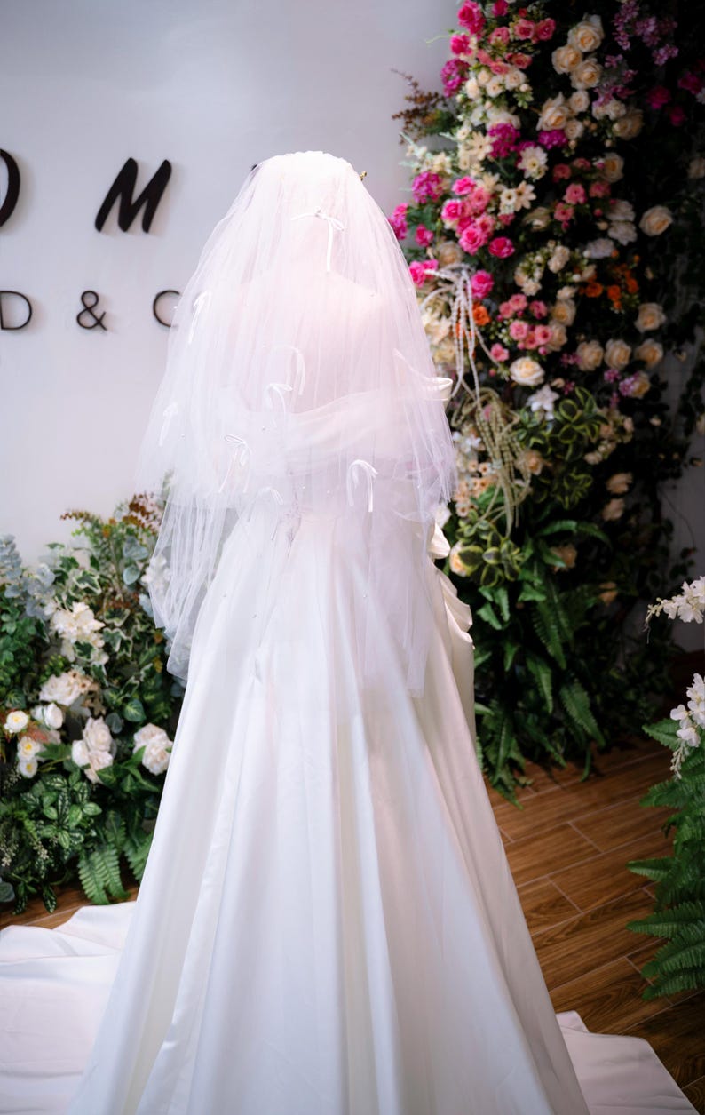 May include: A white wedding dress and veil on a mannequin. The veil is adorned with subtle decorations. The dress has a flowing skirt. The background includes floral arrangements with pink and white flowers, and green foliage.