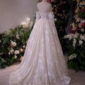 K&ouml;nnte beinhalten: Ein elfenbeinfarbenes Hochzeitskleid mit aufwendigen Spitzendetails. Das Kleid hat lange &Auml;rmel, einen schulterfreien Ausschnitt und einen flie&szlig;enden Rock. Das Kleid wird auf einer Schaufensterpuppe pr&auml;sentiert, mit einem Blumenbogen im Hintergrund.