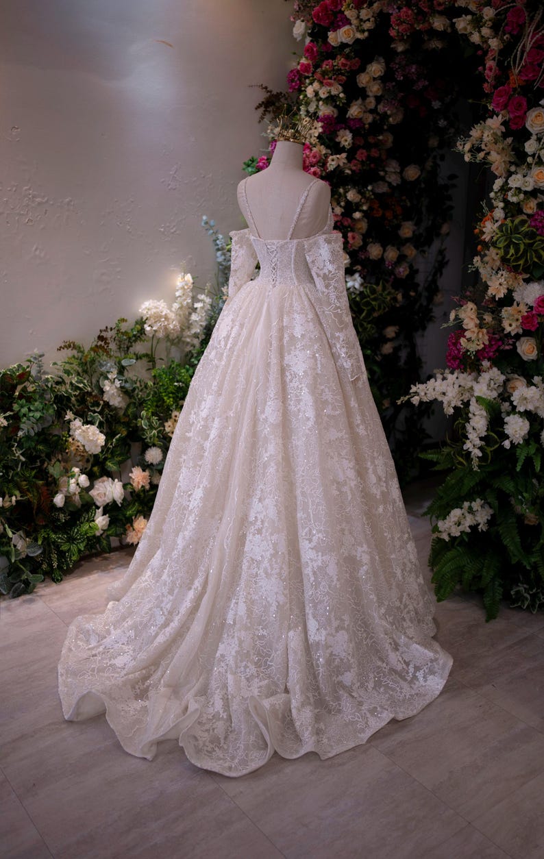K&ouml;nnte beinhalten: Ein elfenbeinfarbenes Hochzeitskleid mit aufwendigen Spitzendetails. Das Kleid hat lange &Auml;rmel, einen schulterfreien Ausschnitt und einen flie&szlig;enden Rock. Das Kleid wird auf einer Schaufensterpuppe pr&auml;sentiert, mit einem Blumenbogen im Hintergrund.
