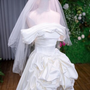 May include: A white satin wedding dress on a mannequin. The off-the-shoulder gown has a ruched bodice and a full skirt with ruffled details. A gold crown and a sheer veil complete the bridal look.