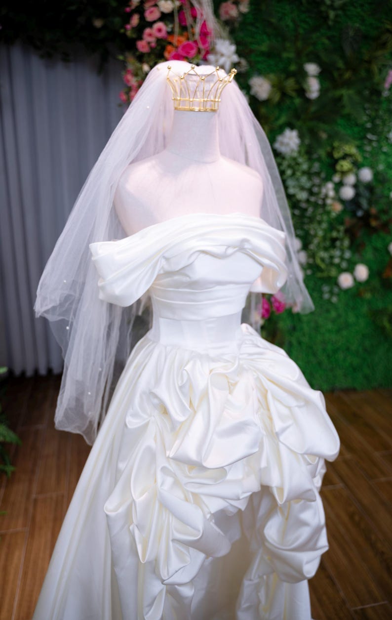 May include: A white satin wedding dress on a mannequin. The off-the-shoulder gown has a ruched bodice and a full skirt with ruffled details. A gold crown and a sheer veil complete the bridal look.