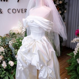May include: A white satin wedding dress on a mannequin, with an off-the-shoulder neckline and a ruffled skirt. A gold crown and veil are on the mannequin's head. The background includes floral arrangements and the text "MY & COVER".