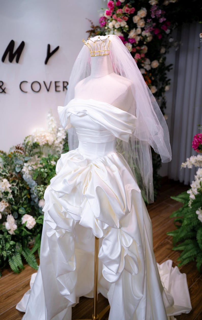May include: A white satin wedding dress on a mannequin, with an off-the-shoulder neckline and a ruffled skirt. A gold crown and veil are on the mannequin's head. The background includes floral arrangements and the text "MY & COVER".