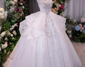 Puede incluir: Vestido de novia blanco adornado, exhibido en un maniquí. El vestido presenta un cuello alto, intrincados bordados y una falda amplia de varias capas. Una corona dorada adorna el maniquí. El fondo incluye un arco floral.