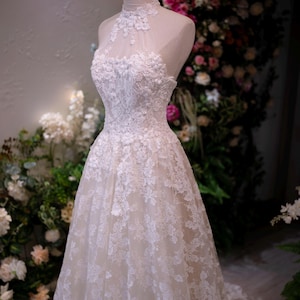May include: A white floor-length gown featuring a high-neck halter bodice with intricate floral lace appliques and a sheer illusion neckline. The dress has a fitted waist that transitions into a full skirt covered in matching white lace embroidery.