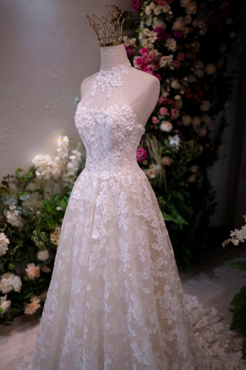 May include: A white floor-length gown featuring a high-neck halter bodice with intricate floral lace appliques and a sheer illusion neckline. The dress has a fitted waist that transitions into a full skirt covered in matching white lace embroidery.