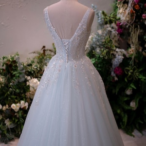 May include: A light blue wedding dress with a V-neckline and a full skirt. The bodice is embellished with sequins and beads. A gold crown sits atop the mannequin head. The dress is displayed against a backdrop of greenery and flowers.