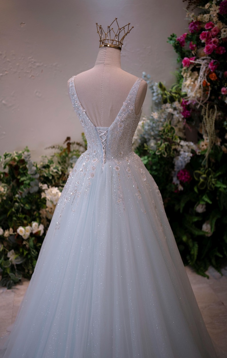 May include: A light blue wedding dress with a V-neckline and a full skirt. The bodice is embellished with sequins and beads. A gold crown sits atop the mannequin head. The dress is displayed against a backdrop of greenery and flowers.