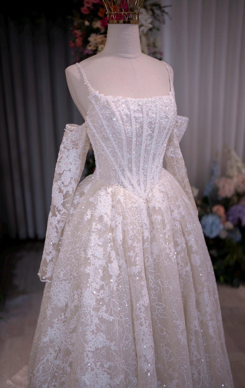 K&ouml;nnte beinhalten: Ein wei&szlig;es Hochzeitskleid mit quadratischem Ausschnitt und schulterfreien &Auml;rmeln. Das Oberteil ist mit vertikalen Paillettenreihen verziert und der Rock weist eine komplizierte Blumenspitze auf. Das Kleid wird auf einer Schaufensterpuppe pr&auml;sentiert und mit einer goldenen Krone gekr&ouml;nt.