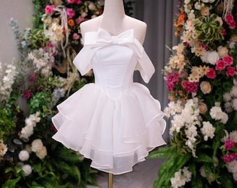 May include: A white strapless mini dress featuring a large bow detail at the neckline, off-the-shoulder sleeves, and a multi-layered ruffled skirt. The dress is displayed on a mannequin wearing a small gold crown on its head.