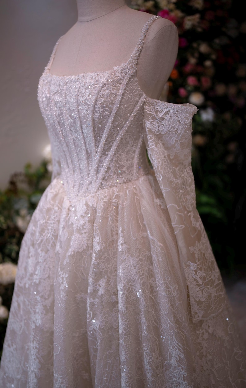 K&ouml;nnte beinhalten: Elfenbeinwei&szlig;es Hochzeitskleid mit aufwendigen Spitzendetails und funkelnden Verzierungen. Das Oberteil hat ein strukturiertes Design mit quadratischem Ausschnitt und schulterfreien &Auml;rmeln. Der Rock flie&szlig;t anmutig und schafft eine romantische und elegante Silhouette.