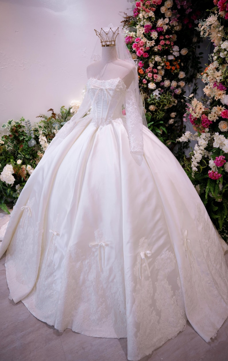 May include: A white ball gown with a corset-style bodice, long lace sleeves, and a full skirt featuring lace appliques and small ribbon bows. The dress is paired with a sheer veil and a gold-toned crown headpiece resting on the mannequin.