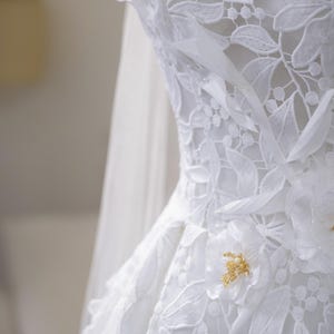 May include: Close-up of a white wedding dress with detailed floral lace. The dress has delicate flower embellishments with gold bead centres. A sheer veil is partially visible, adding to the elegant bridal design.