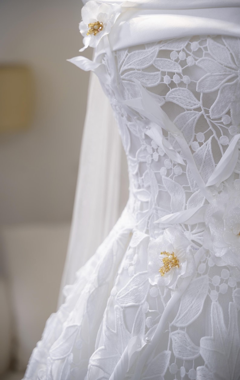 May include: Close-up of a white wedding dress with detailed floral lace. The dress has delicate flower embellishments with gold bead centres. A sheer veil is partially visible, adding to the elegant bridal design.