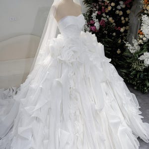 Könnte beinhalten: Ein trägerloses weißes Brautkleid auf einer Schaufensterpuppe, mit einem geschichteten, gerüschten Rock und einer langen Schleppe. Eine goldene Krone und ein Schleier befinden sich auf dem Kopf der Schaufensterpuppe. Im Hintergrund ein Blumenarrangement und der Text "OMY ED & COVER".