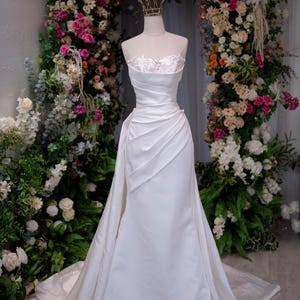 May include: A strapless white wedding gown on a mannequin, featuring a fitted bodice with lace detailing and a draped skirt that flows to the floor. A gold crown sits atop the mannequin's head. The background is a floral archway.