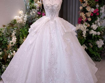 Puede incluir: Un vestido de gala blanco con escote corazón, un corpiño ilusión de cuello alto con intrincados abalorios plateados y una falda voluminosa con paneles de tela superpuestos. El vestido está adornado con lentejuelas brillantes y una corona dorada.