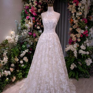 May include: A floor-length white lace gown featuring a high halter neckline with intricate floral embroidery, a fitted bodice, and a full A-line skirt. The dress is displayed on a mannequin wearing a small gold crown on the neck area.