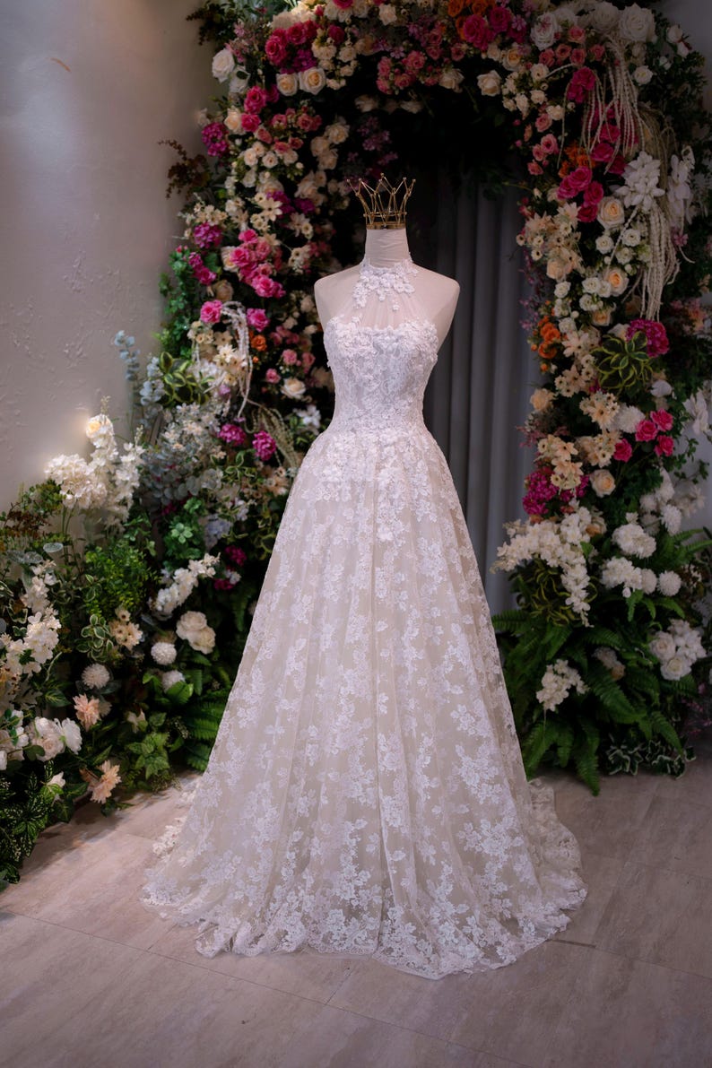 May include: A floor-length white lace gown featuring a high halter neckline with intricate floral embroidery, a fitted bodice, and a full A-line skirt. The dress is displayed on a mannequin wearing a small gold crown on the neck area.