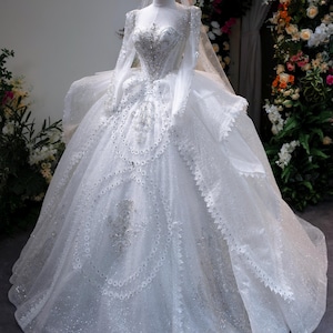 Könnte beinhalten: Ein weißes Hochzeitskleid mit langen Ärmeln und einem weiten Rock, verziert mit aufwendiger Perlenarbeit und glitzernden Details. Das Kleid wird auf einer Schaufensterpuppe mit goldener Krone und Schleier präsentiert. Ein klassisches Brautkleid.