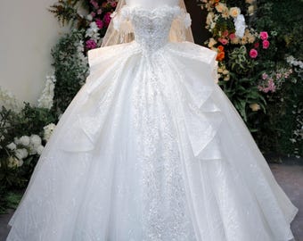 Puede incluir: Un vestido de novia blanco estilo corte de baile con escote de hombros caídos, intrincados bordados de lentejuelas plateadas y una falda voluminosa con volantes en capas. Se acompaña de un velo transparente y una pequeña corona dorada.