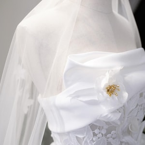 May include: A white wedding dress on a mannequin. The strapless bodice features floral lace, a satin bow, and a flower with gold accents. A sheer veil drapes over the dress, creating an elegant look.