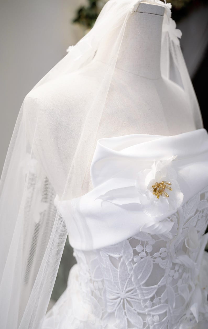 May include: A white wedding dress on a mannequin. The strapless bodice features floral lace, a satin bow, and a flower with gold accents. A sheer veil drapes over the dress, creating an elegant look.