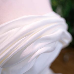 May include: Close-up of white draped fabric, possibly silk or satin, with soft folds and subtle highlights. The fabric appears to be part of a garment, with a blurred background suggesting a wedding dress or formal wear.