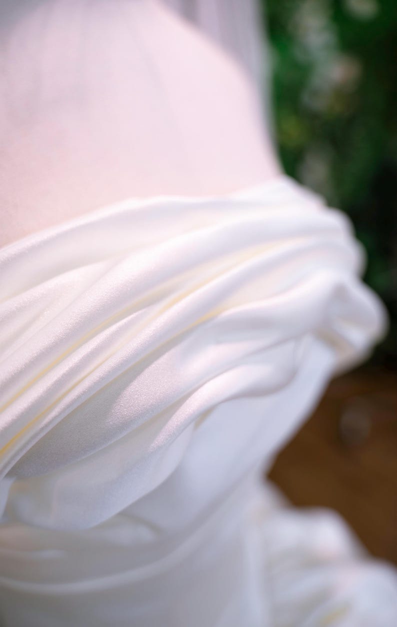 May include: Close-up of white draped fabric, possibly silk or satin, with soft folds and subtle highlights. The fabric appears to be part of a garment, with a blurred background suggesting a wedding dress or formal wear.