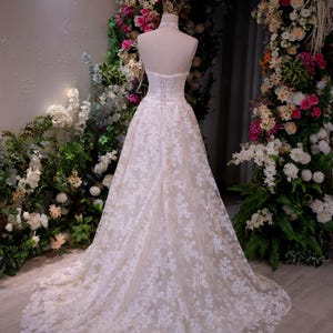 May include: A floor-length, strapless white gown featuring an intricate lace overlay with a floral pattern, a corset-style bodice with lace-up back detailing, and a full, flowing skirt that extends into a train, displayed on a mannequin.