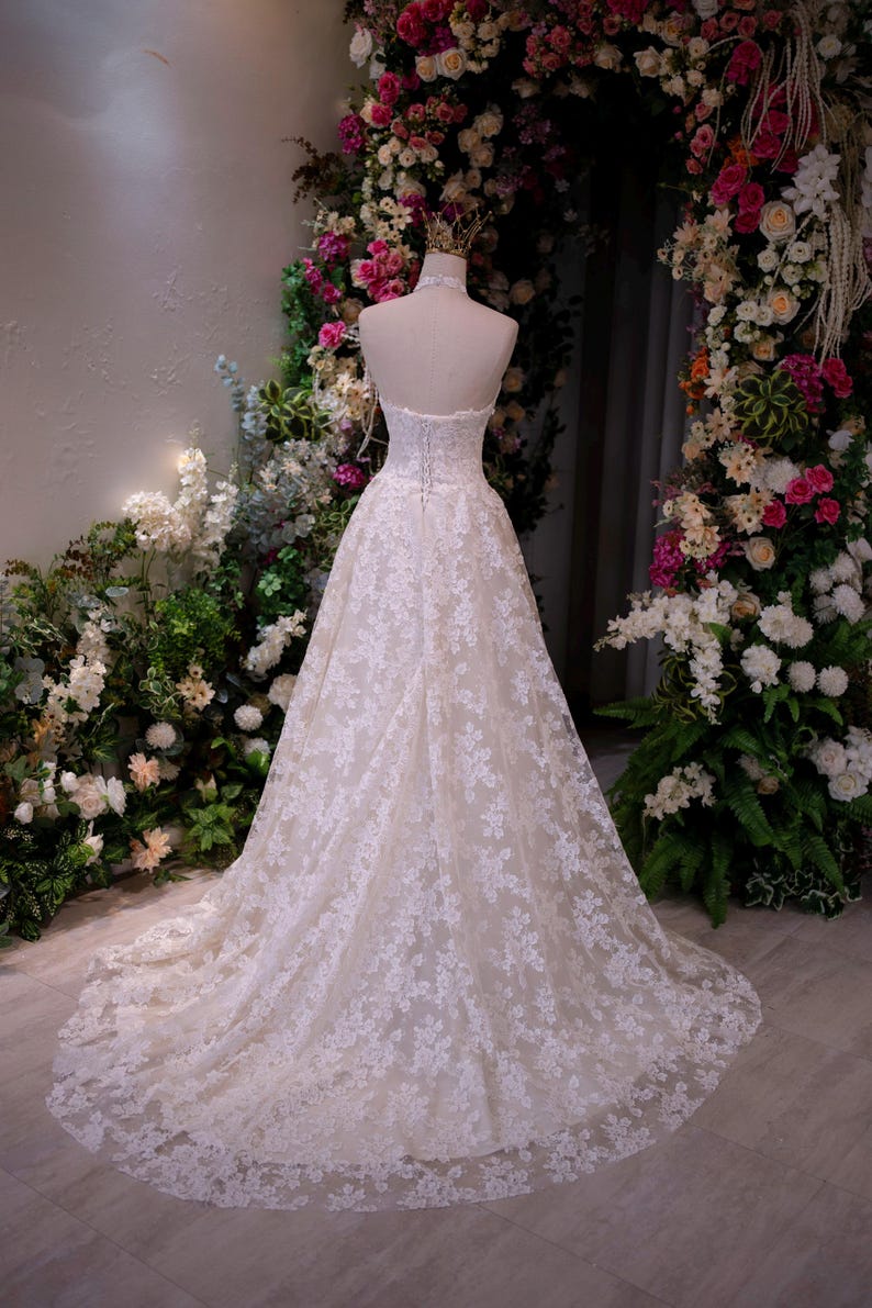 May include: A floor-length, strapless white gown featuring an intricate lace overlay with a floral pattern, a corset-style bodice with lace-up back detailing, and a full, flowing skirt that extends into a train, displayed on a mannequin.
