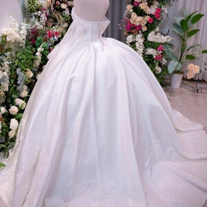 May include: A white ball gown dress with an off-the-shoulder neckline, a corset-style bodice, and a voluminous skirt featuring lace detailing along the hemline. The dress is displayed on a mannequin wearing a gold-toned crown headpiece.