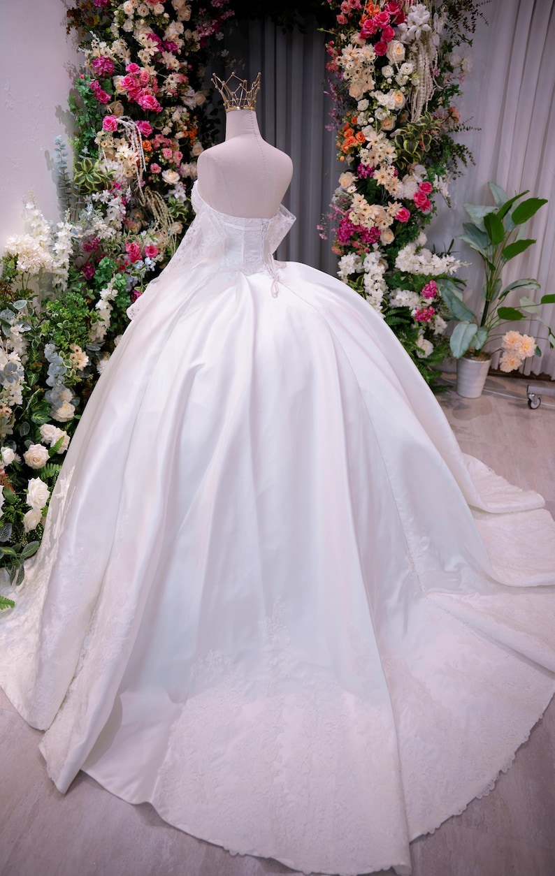 May include: A white ball gown dress with an off-the-shoulder neckline, a corset-style bodice, and a voluminous skirt featuring lace detailing along the hemline. The dress is displayed on a mannequin wearing a gold-toned crown headpiece.